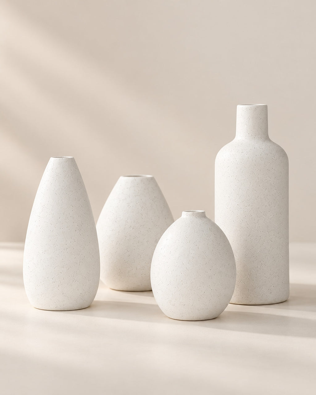 Four matte white ceramic bud vases grouped on a warm off-white surface.