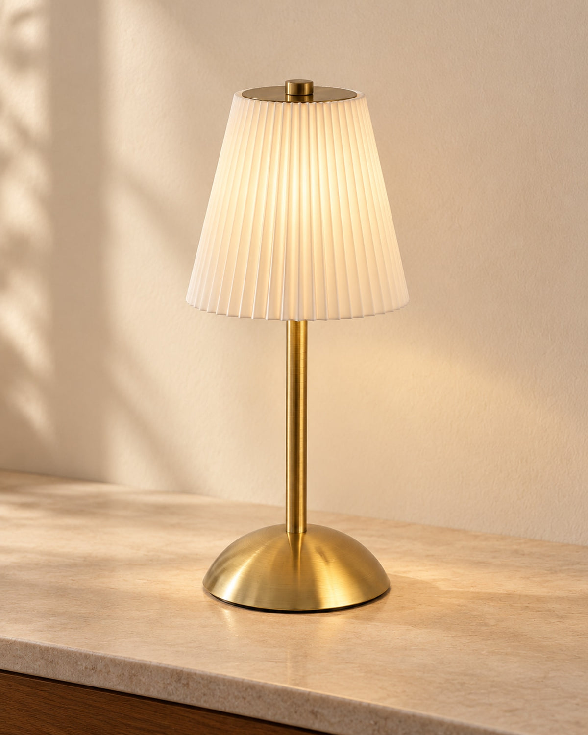 Cordless table lamp with a white pleated shade and brass-coloured stem, switched on.