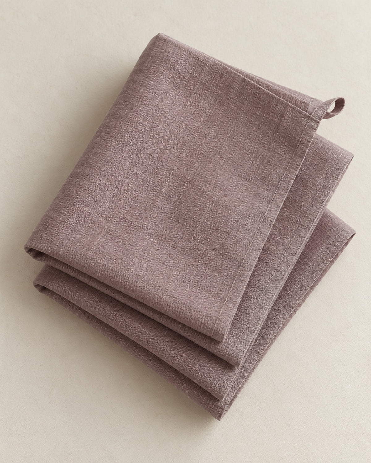 Three folded dusty taupe linen tea towels on a warm off-white surface.
