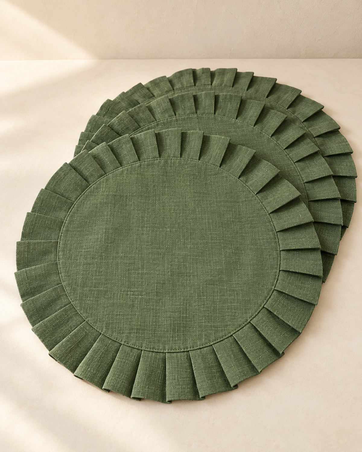 Round green linen placemat with a pleated ruffle edge on a warm off-white surface.