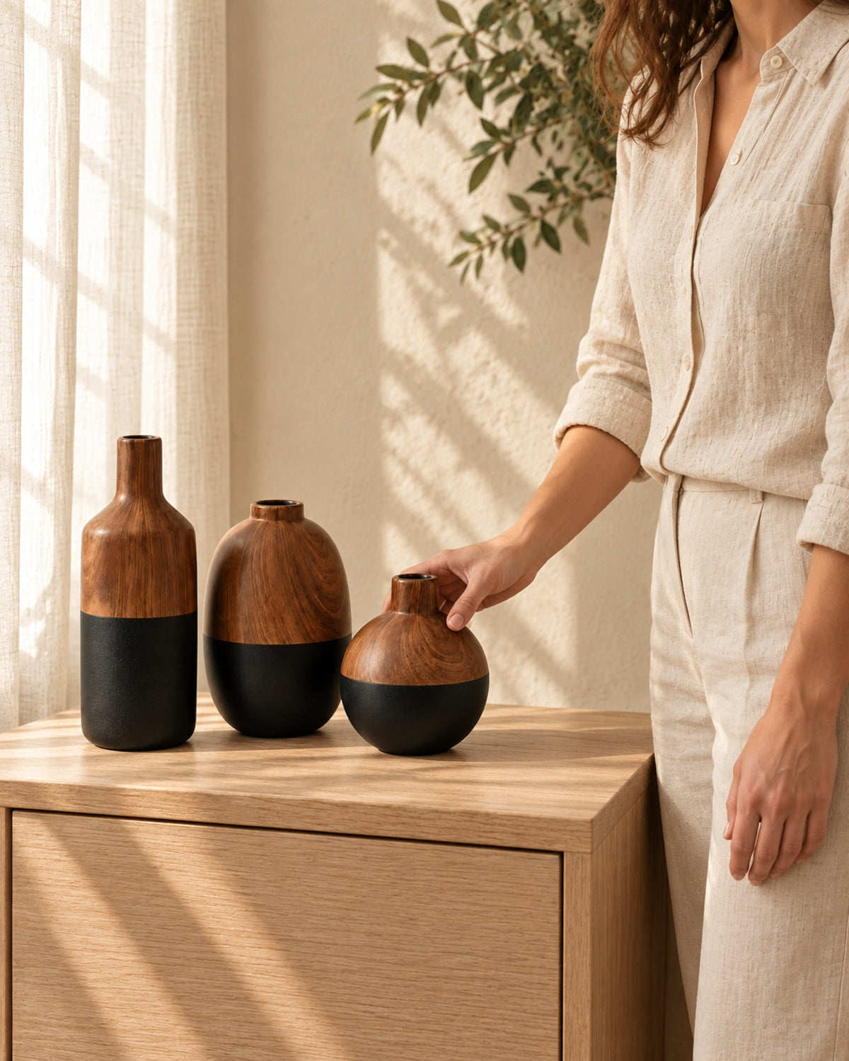 A person in linen placing the round walnut and black vase next to the bottle and oval vase on an oak cabinet.
