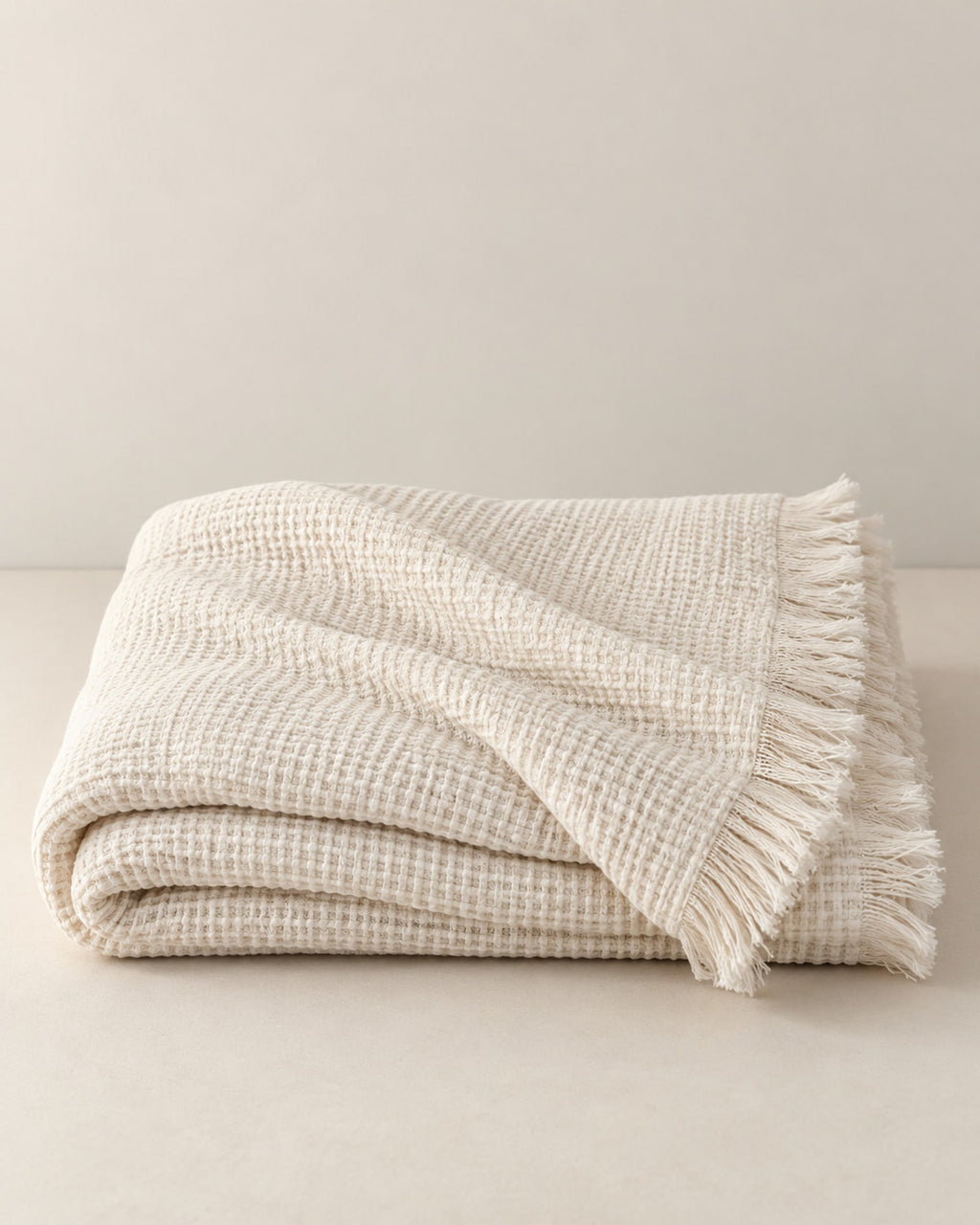 Folded cream white waffle cotton throw with fringe on a warm off-white surface.