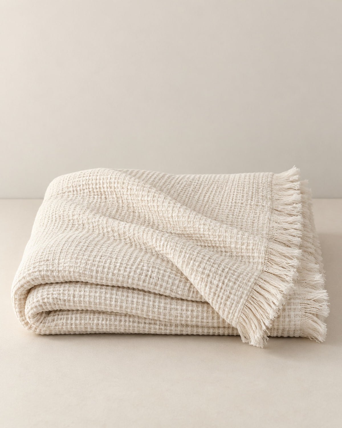 Folded cream white waffle cotton throw with fringe on a warm off-white surface.